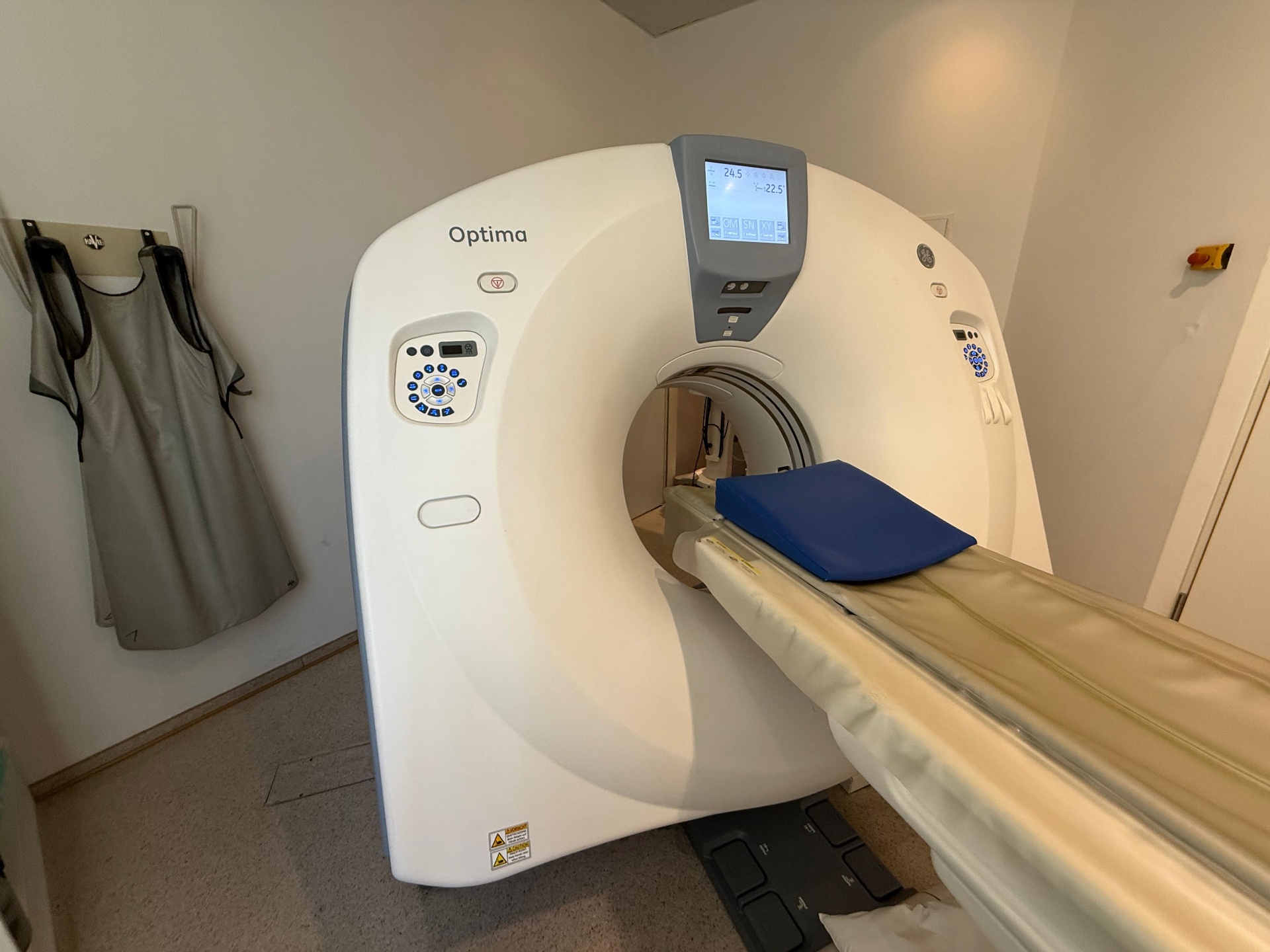 GE Optima CT520 angled gantry and patient table view with operator keypad and lead apron visible in scan room